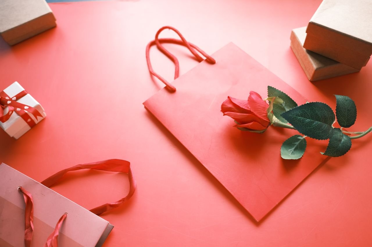 A romantic flat lay featuring a red rose on a paper bag, gift boxes, and a pink background.