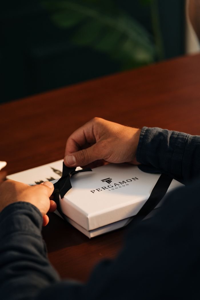 Person unwrapping a Pergamon gift box on a wooden table, creating a cozy indoor atmosphere.