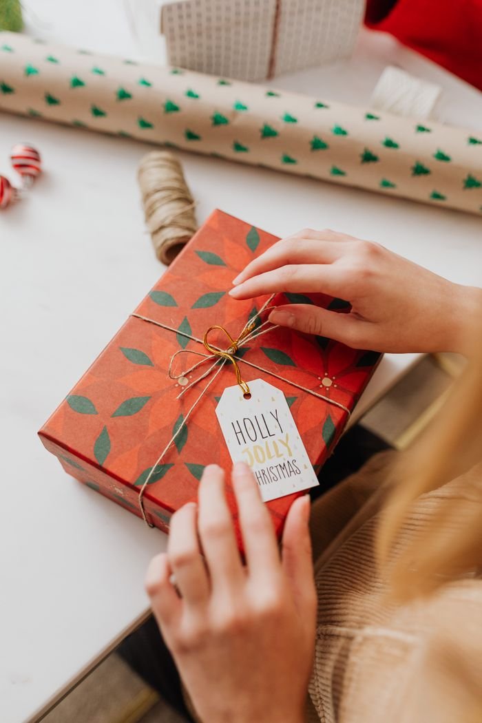 heros-img Hands wrapping a festive Christmas gift in red paper with a 'Holly Jolly Christmas' tag.